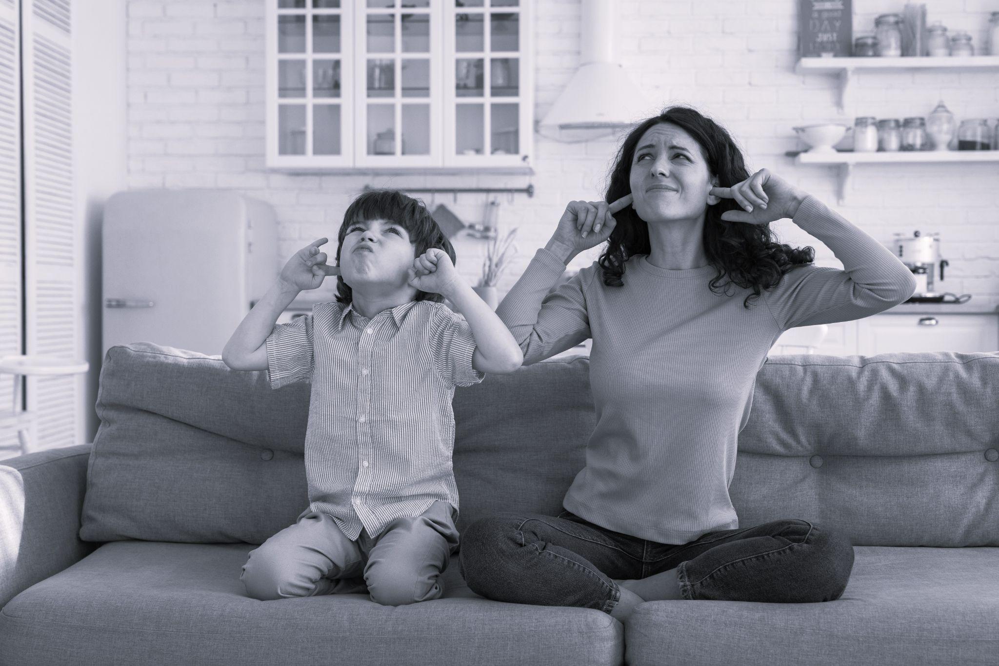 Frowning mom and son sit on couch covering ears from noisy music