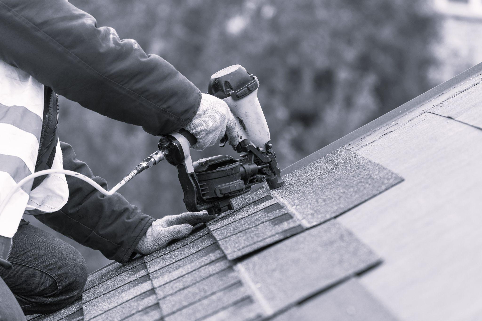 Roofer worker in uniform work wear using air gun and installing asphalt top of the roof