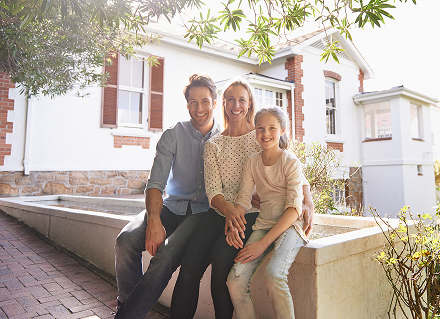 A family of three sits happily on a stone ledge in front of a charming, sunlit brick house. They are smiling, conveying warmth and contentment.