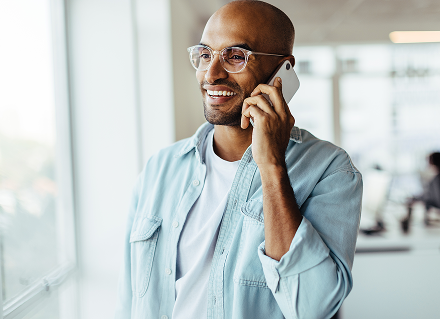A man in glasses and a light blue shirt smiles as he talks on a phone by a bright window in an office.