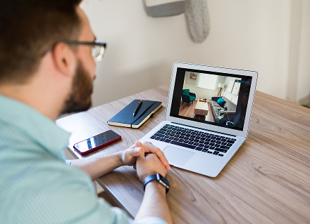 A man with glasses sits at a wooden desk, viewing a room layout on a laptop. Nearby are a notebook and smartphone, conveying a focused and professional atmosphere.